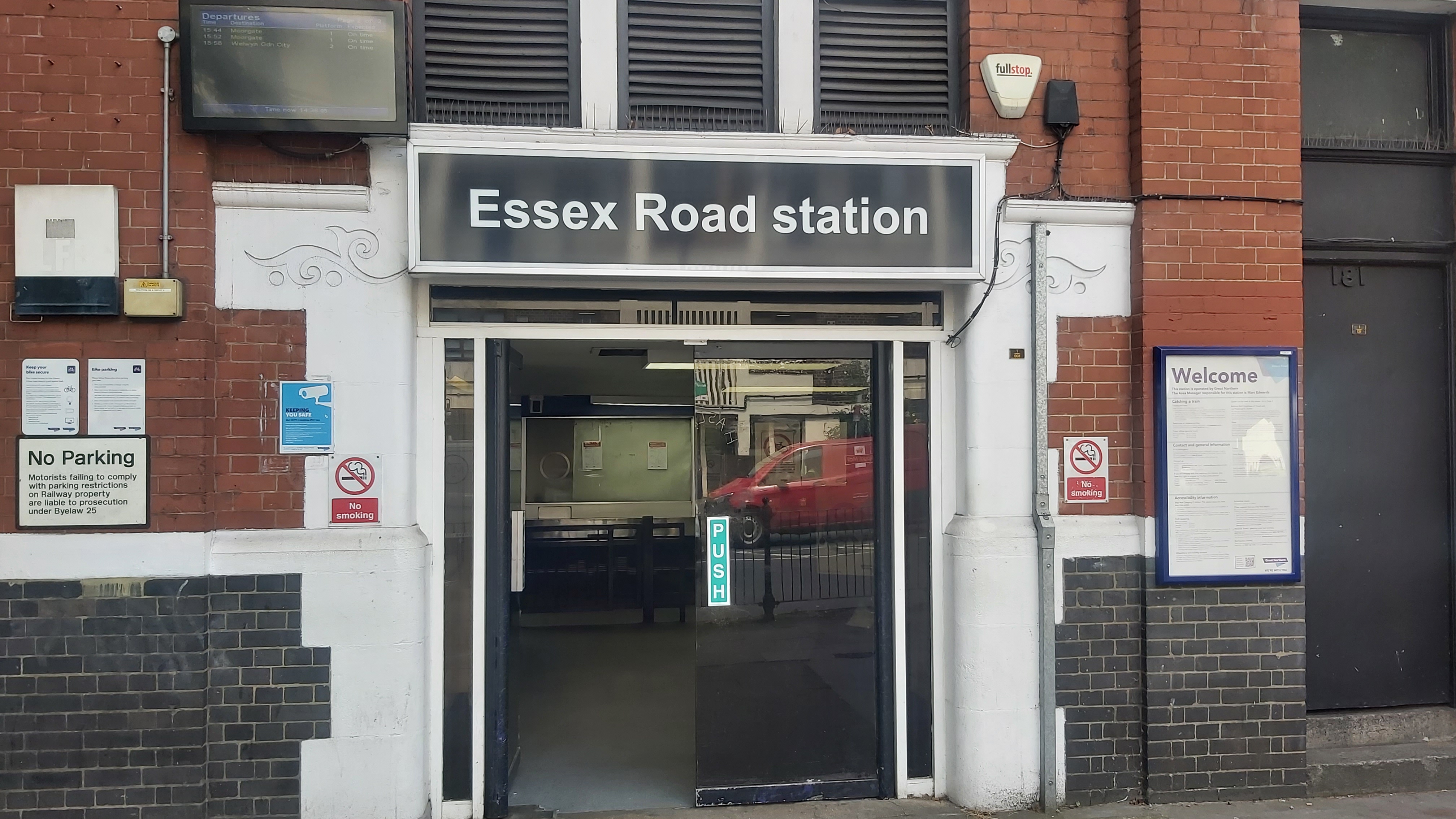 The exterior of Essex Road station. Visible is a variety of notices and posters, a large 'Essex Road' sign, and a TV displaying the next trains. Two 'No Smoking'
    signs flank the double-door entrance. A Royal Mail van is reflected onto the doors.